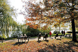 2010 Die Herbstsonne scheint durch das Laub der Bume am Alsterufer von Hamburg Harvestehude - Spaziergnger gehen auf dem Alsterweg - andere Besucher der Grnanlage sitzen auf den Ruhebnken am Ufer der Aussenalster.