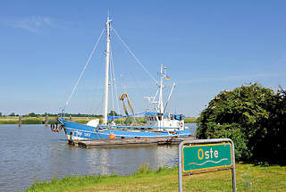 5106 Schild Oste - grner Grund mit weisser Schrift an der Oste bei Neuhaus - ein blauer Fischkutter liegt am Ufer.